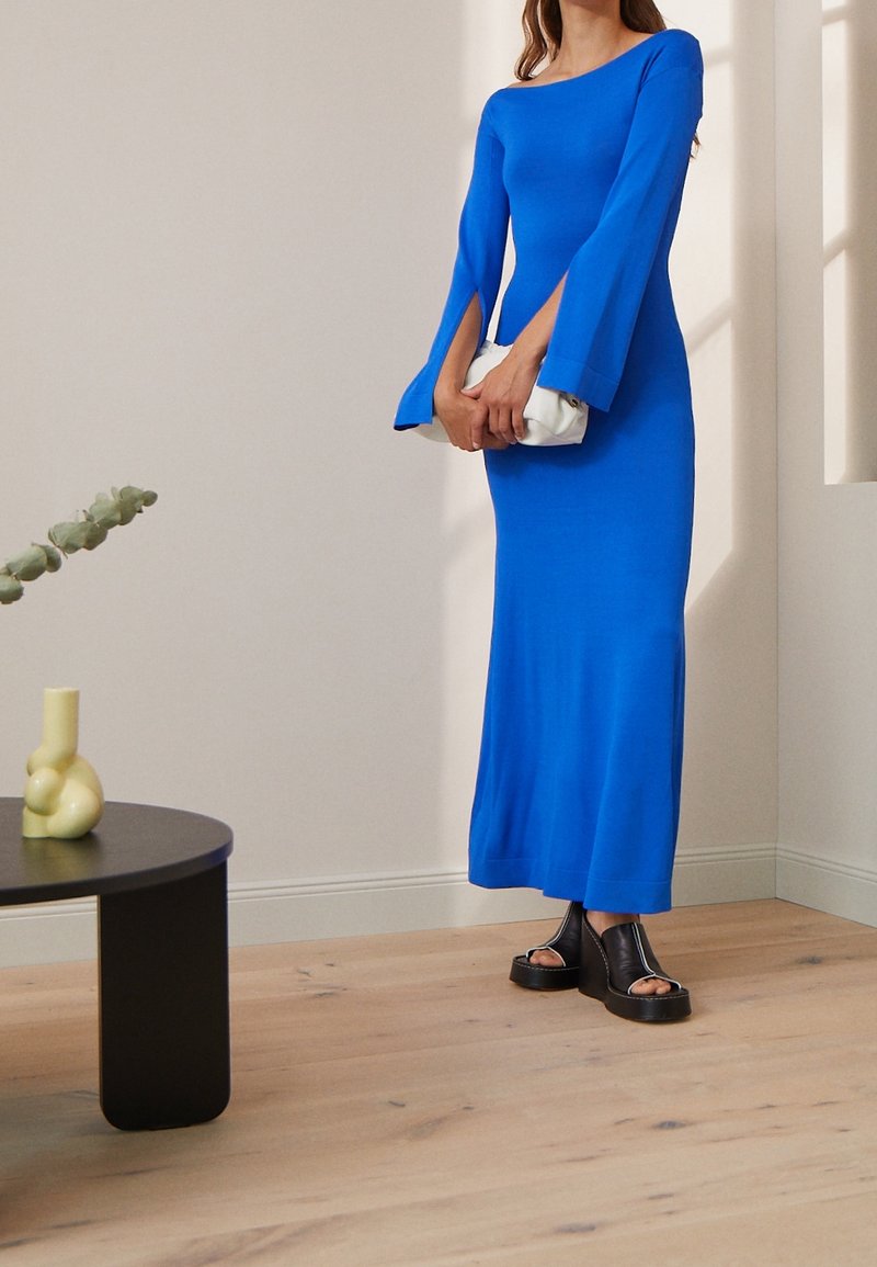 Blå långärmad klänning med utställda ärmar, slim fit, texturerat material. Bärs med svarta plattformssandaler och en vit clutch. Minimalistisk bakgrund.