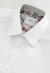 Vit skjorta med knappar i en texturerad tyg, med en krage inuti med blommönster och en etikett som visar "NEXT TAILORING."