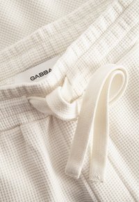 Crèmekleurige gestructureerde stof met een rasterpatroon, voorzien van een trekkoord en een label met de tekst "GABBA." Zacht, lichtgewicht materiaal.