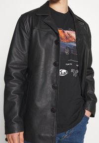 Svart läderjacka med krage, med fem knappar och en slät yta. Buren över en svart tryckt t-shirt och blå jeans.