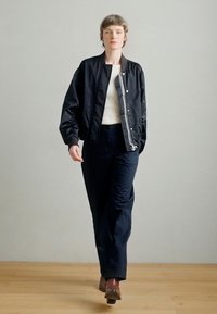 Jachetă bomber neagră cu finisaj satinat și manșete tricotate, asortată cu pantaloni închiși la culoare și pantofi maro, pe podea de lemn, cu un fundal neutru.