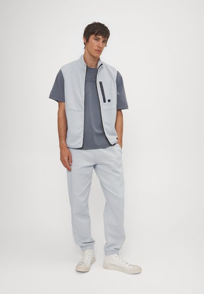 Lysblå ærmeløs zip-vest over en navy t-shirt med korte ærmer, parret med matchende lysblå joggers og hvide sneakers. Glat stofstruktur.