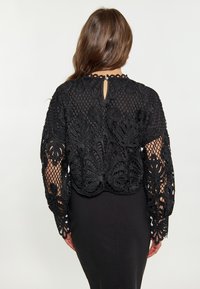Top corto in pizzo nero con ricami floreali, maniche lunghe, schiena aperta con dettaglio a bottone, abbinato a un vestito aderente nero. Texture liscia.
