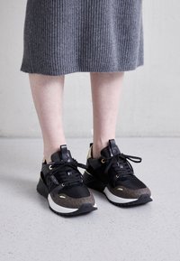 Pies que llevan zapatillas de diseñador negras y marrones con acentos dorados, de pie sobre un suelo de color gris claro salpicado, visibles desde la mitad de la pantorrilla hacia abajo.