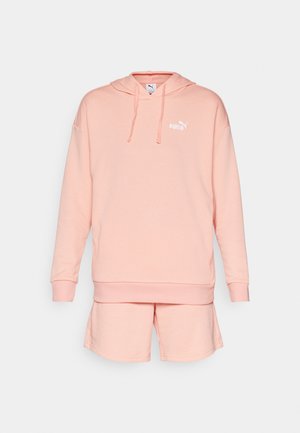 Sudadera de color melocotón con un bolsillo canguro y capucha con cordón, combinada con pantalones cortos a juego. La tela parece suave y tiene un logo minimalista.