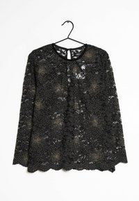 Top en dentelle noire à manches longues avec des motifs à pois, décolleté rond et ourlet en forme de vagues. Suspendu à un crochet noir sur fond blanc.