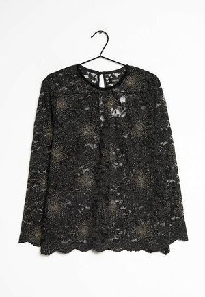 Top lungo a maniche in pizzo nero con motivi a pois, scollo rotondo e orlo ondulato. Appeso a un gancio nero su uno sfondo bianco.