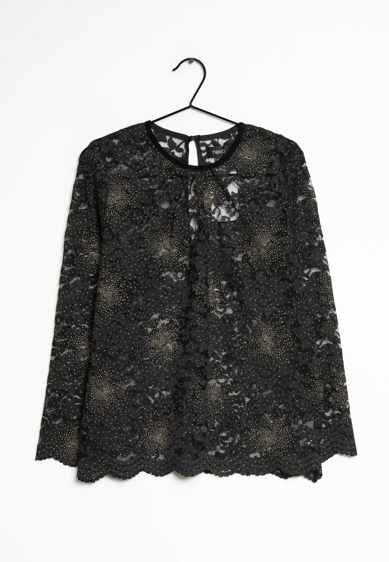 Top en dentelle noire à manches longues avec des motifs à pois, décolleté rond et ourlet en forme de vagues. Suspendu à un crochet noir sur fond blanc.