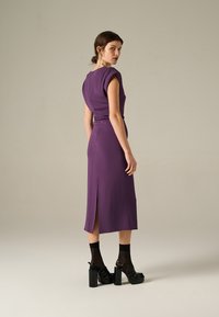 Abito midi viola con maniche a cappuccio, vita cintrata e dettaglio con spacco sul retro. Indossato con scarpe platform nere e calze trasparenti.