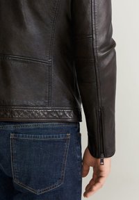 Hnědá kožená bunda s texturovaným povrchem, vybavená zipem na manžetě a detaily švů. Kombinována s tmavě modrými džínami.