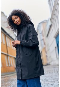 Veste à capuche noire imperméable avec des poignets élastiques, des poches latérales et une coupe décontractée. Portée sur un jean large bleu dans une rue en pavés.