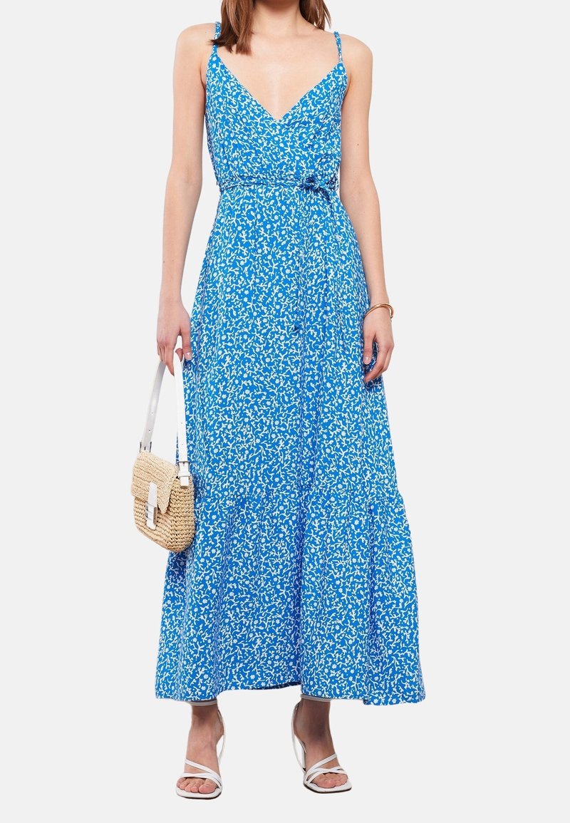 Blauwe bloemen maxi jurk met spaghettibandjes, een V-halslijn en een gelaagde rok. Draagt een paar witte sandalen en een gestructureerde beige handtas.