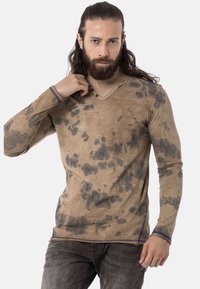 Langärmliges, V-Ausschnitt-Shirt aus strukturiertem beigefarbenem Stoff mit schwarzen Spritzmustern. Verfügt über blaue Nähte entlang dem Saum und dem Ausschnitt.