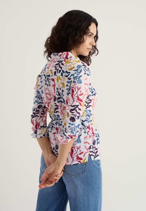 Moteris su tamsiais garbanotais plaukais, dėvinti ilgomis rankovėmis gėlių raštu margintą marškinių švarkelį ir mėlynas džinsus, stovinti sukryžiavusi rankas už nugaros.