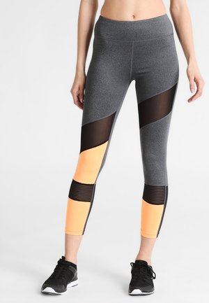 Kvinde iført grå leggings med sorte mesh- og orange paneler, kombineret med sorte sportssko, stående mod en ensfarvet baggrund.