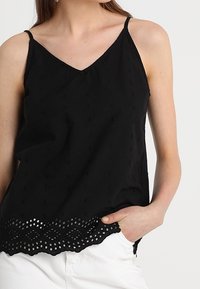 Black sleeveless top with a V-neck, featuring textured fabric and a decorative lace trim at the hem with circular cut-outs.