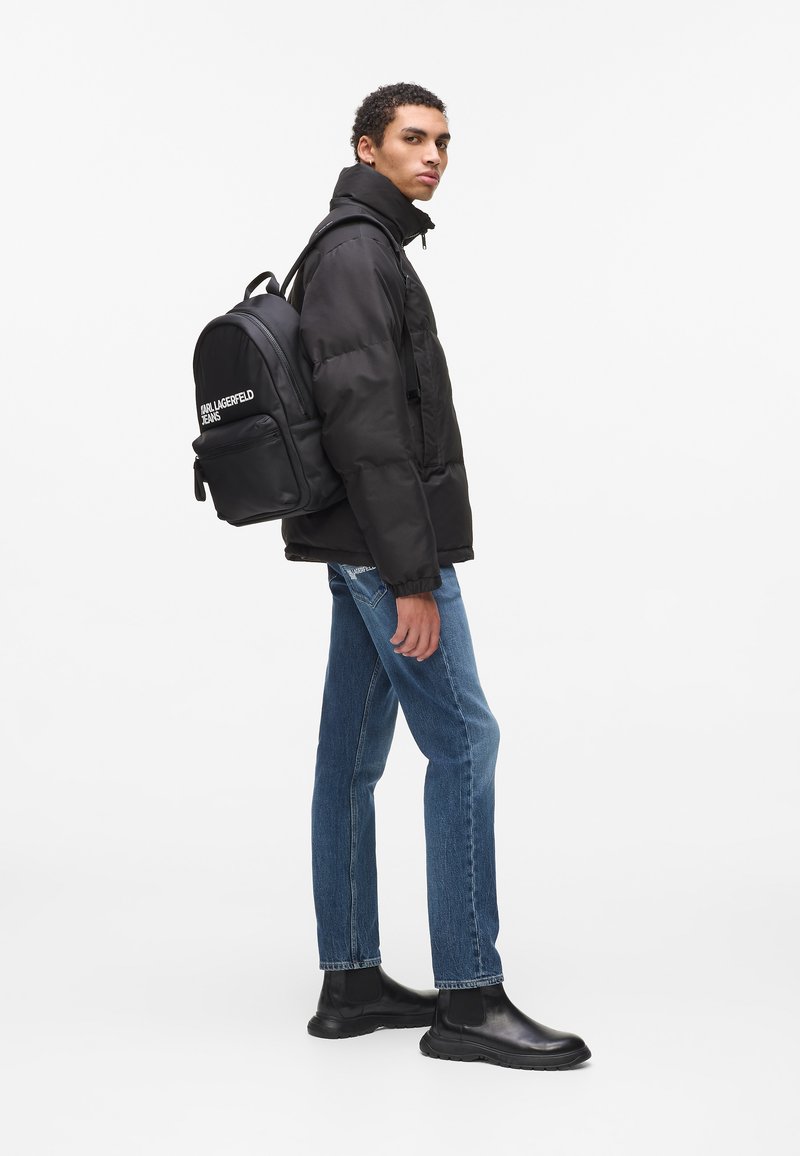 Μαύρο παλτό με επένδυση πάνω από λευκό πουκάμισο, μπλε τζιν και μαύρες Τσέλσι μπότες. Μαύρο σακίδιο πλάτης με λευκό λογότυπο "Karl Lagerfeld Jeans".