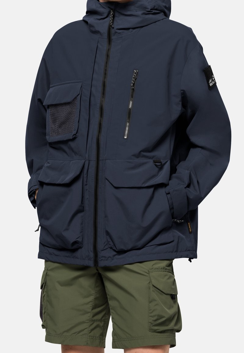 Personne portant une veste à capuche bleu marine avec plusieurs poches et un short cargo vert olive, les mains partiellement dans les poches.