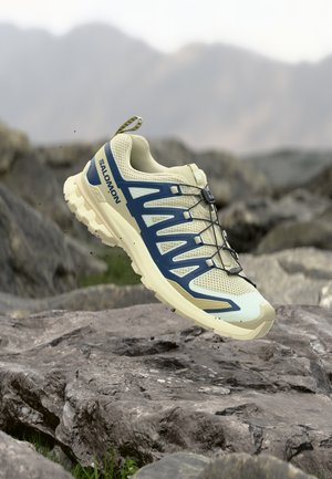 Lichtbeige en blauwe Salomon trailrunningschoen zwevend boven rotsachtig terrein met een vervaagde bergachtergrond.