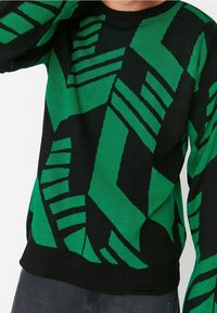 Pull noir avec des motifs géométriques verts, comprenant des bandes et des formes angulaires. Col et poignets côtelés. Tissu tricoté léger.