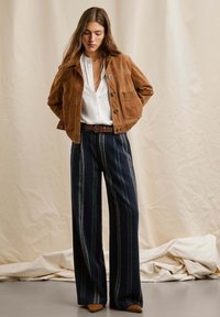 Veste en suede marron avec un col, quatre poches à l'avant et des boutons. Chemise blanche en dessous. Pantalons larges rayés bleu marine avec une ceinture marron.