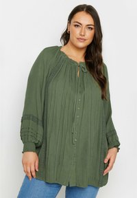 Grön knäppblus med texturerad tyg, elastiska ärmar, volangkrage och spetsdetaljer längs fram och ärmar.