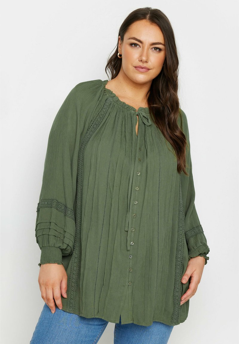 Grön knäppblus med texturerad tyg, elastiska ärmar, volangkrage och spetsdetaljer längs fram och ärmar.