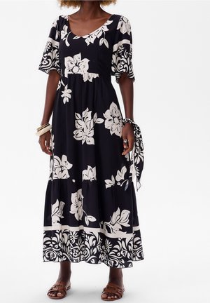 Frau trägt ein schwarzes Maxikleid mit großen weißen Blumenmustern, mit flatternden Ärmeln, Sandalen und Armbändern vor weißem Hintergrund.