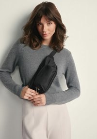 Czarna nylonowa torba na ramię o gładkiej fakturze, z zamykaną na zamek kieszenią i regulowanym paskiem. Nosi się ją na szarym swetrze.