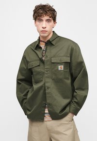 Πουκάμισο μακρυμάνικο σε ελιά χρώμα με κουμπιά μπροστά, δύο τσέπες στο στήθος και λογότυπο. Η υφή είναι απαλή και το ύφασμα φαίνεται ανθεκτικό.