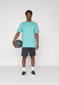Camiseta deportiva turquesa con un pequeño logo verde, combinada con pantalones cortos negros. El jugador sostiene un balón de baloncesto, vistiendo zapatillas negras y calcetines blancos.