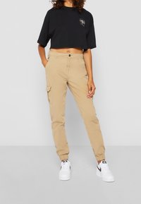 Pantalons cargo beige avec des poignets élastiques et des poches latérales, associés à un t-shirt noir court avec un emblème graphique. Baskets blanches avec des accents noirs.