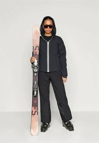 Donna in abbigliamento da sci nero, occhiali da sole e stivali, tiene uno sci rosa e nero in posizione verticale contro uno sfondo semplice.