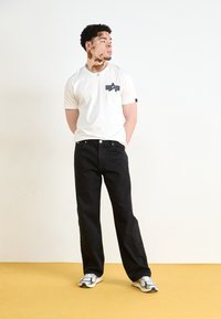 Kremfarget t-skjorte med svart grafikkdetalj, parret med svarte vide jeans med subtil broderi, og hvite sneakers med svarte aksenter.