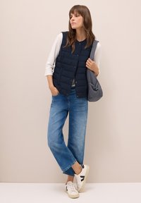 Donna che indossa un gilet trapuntato blu scuro, una maglietta bianca a maniche lunghe, jeans a gamba larga, sneakers bianche e porta una borsa a tracolla grigia, in piedi davanti a un muro beige.