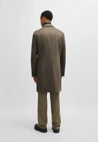 Cappotto di lana marrone con taglio dritto, colletto a punte, spacco posteriore e polsini con bottone, abbinato a pantaloni beige e scarpe nere.