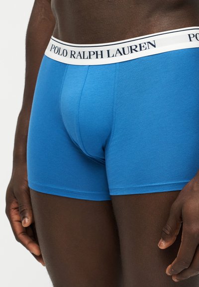 Άνδρας που φοράει λαμπερό μπλε boxer Polo Ralph Lauren με λευκή ζώνη και μαύρο λογότυπο, στέκεται με τα χέρια χαλαρά δίπλα στο σώμα.
