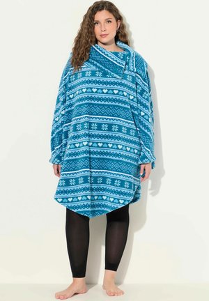 Poncho in fleece turchese con motivi geometrici bianchi, inclusi cuori e fiocchi di neve; design oversize con un grande colletto. Leggings neri indossati sotto.