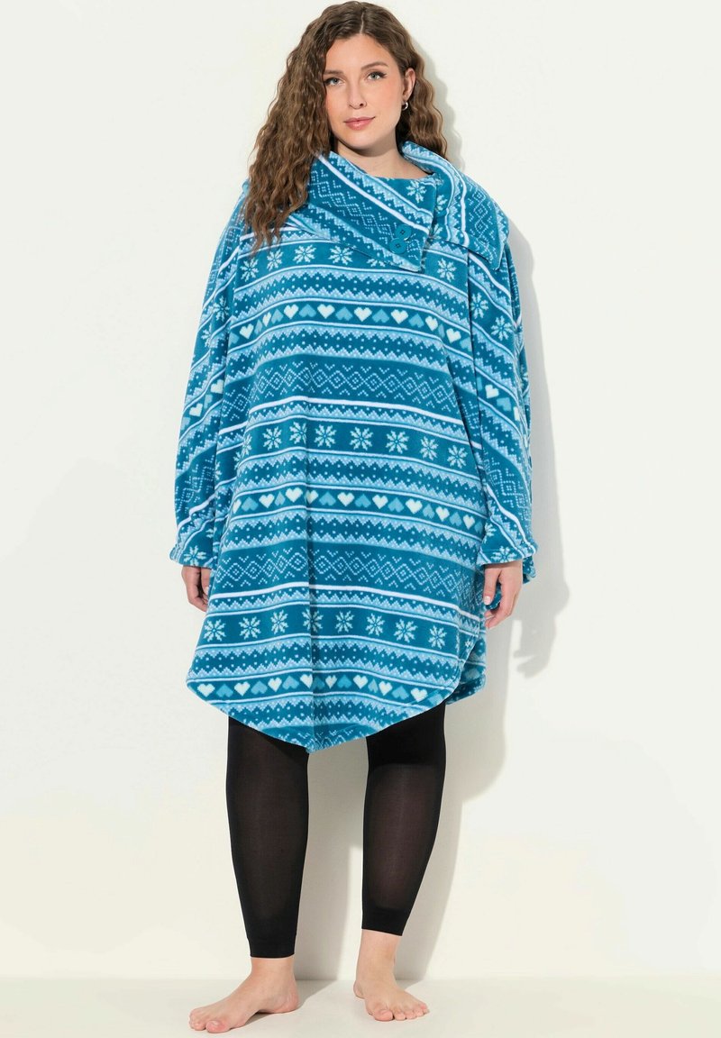 Poncho in fleece turchese con motivi geometrici bianchi, inclusi cuori e fiocchi di neve; design oversize con un grande colletto. Leggings neri indossati sotto.