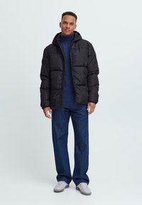 Geacă pufină neagră cu glugă, având un design matlasat, purtată peste un pulover bleu și asociată cu blugi închisi și adidași deschiși.