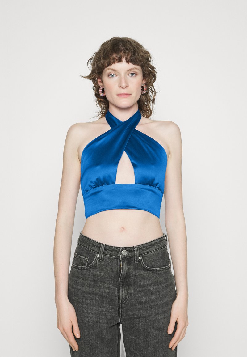 Top cropped de cetim azul em halter com um recorte em formato de chave e alças cruzadas, combinado com jeans pretos de cintura alta. Textura suave e design ajustado.