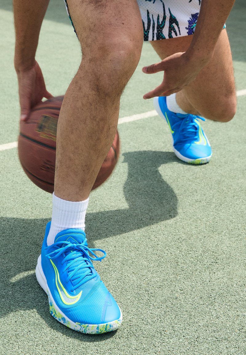 Modré sportovní boty s texturovaným svrškem a vzorovanou podrážkou. Design obsahuje jasně žluté akcenty a šňůrky. Basketbal je v pohybu.