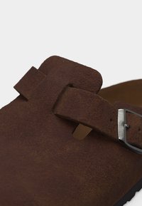 Κοντινό πλάνο καφέ σουέντ παπουτσιού χωρίς κορδόνια με ανοιχτή μύτη, ρυθμιζόμενο λουράκι και ασημένια πόρπη σε λευκό φόντο.