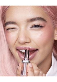Rouge à lèvres dans un tube transparent avec des accents métalliques, appliqué sur des lèvres teintées de rose clair. Le mannequin a des cheveux longs roses et un teint pâle.