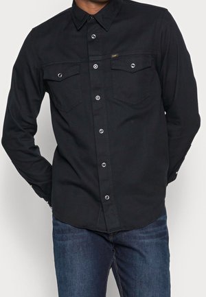 Homme portant une chemise noire boutonnée avec poches à rabat et un jean bleu, mains positionnées derrière le dos.