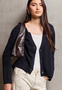 Granatowy sweter z dzianiny z przodu zapinany na guziki, noszony na białej bluzce. Modelka trzyma ciemnobrązową skórzaną torbę z plecionym detalem.