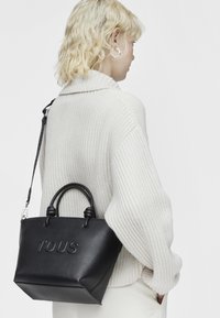 Borsa tote in pelle nera con manici annodati, caratterizzata dal logo "TOUS" in rilievo, posizionata all'anca di una persona in un maglione crema.