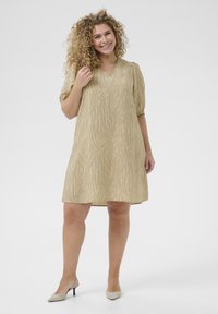 Vestido beige con un patrón abstracto texturizado, con mangas cortas abullonadas y escote en V, combinado con tacones de color claro.