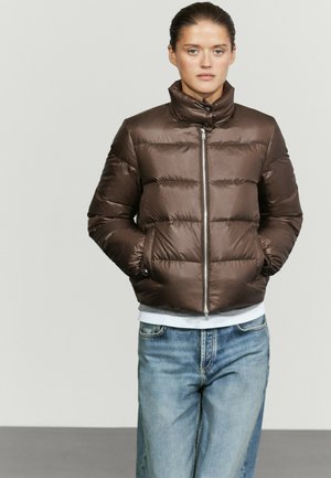 Junge Frau trägt eine braune Steppjacke mit Händen in den Taschen und blaue Jeans, steht vor einem schlichten hellen Hintergrund.