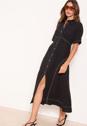 Frau trägt ein schwarzes, knöpfbares Midikleid mit weißen Nähten, orange getönte Sonnenbrille und schwarze Sandalen, steht an einer weißen Wand.